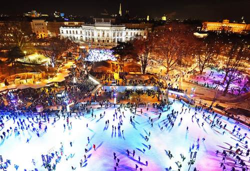 Großaufnahme des Wiener Eistraums samt Marktständen, einer wunderschön beleuchteten Eisfläche und vielen BesucherInnen. Hinten nimmt das festliche Wiener Rathaus das Bild ein.